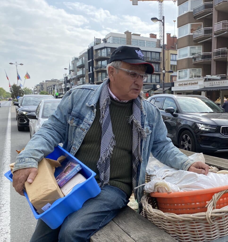 Un homme portant un foulard, une veste en jeans et un pull vert assis sur une charrette en bois, avec un panier en osier à côté de lui