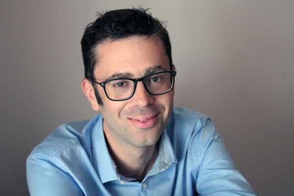 Photo de Nicolas Boubou cadrée aux épaules. Il porte des lunettes et une chemise bleu ciel, et il se tient devant un fond gris unis