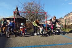 7 enfants sur des petits vélos, accompagnés de deux animatrices dans une cour d'école devant un goal