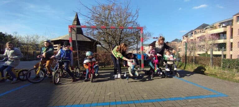 7 enfants sur des petits vélos, accompagnés de deux animatrices dans une cour d'école devant un goal