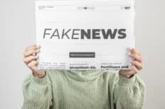 Une femme au pull vert cachant sa tête avec un journal sur lequel est écrit "Fake News"