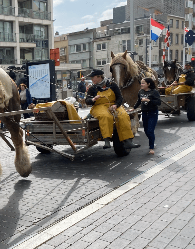 Une femme en salopette jaune avec des bottes en caoutchouc, assise sur une charrette en bois, avec un cheval qui la traine et une petite fille qui marche à côté