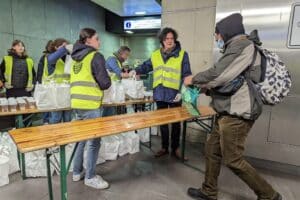 Bénévoles portant un gilet jaune fluo distribuant des sacs blancs de repas. Une personne vient en chercher un