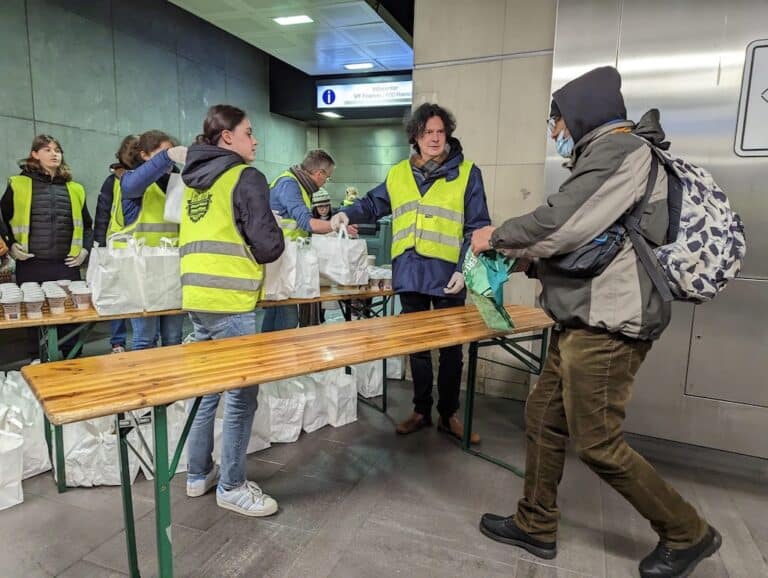 Bénévoles portant un gilet jaune fluo distribuant des sacs blancs de repas. Une personne vient en chercher un