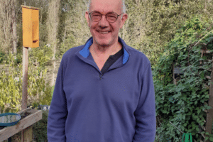 Philippe Hensmans, dehors devant des arbres, les mains dans les poches et souriant à la caméra