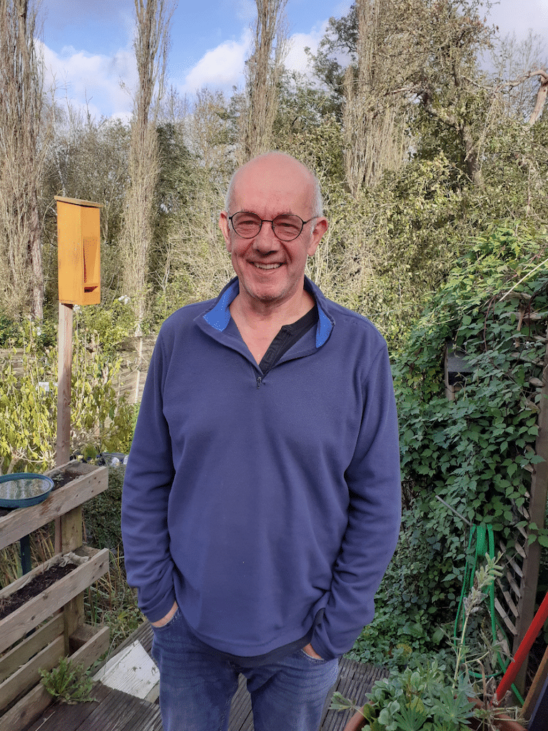 Philippe Hensmans, dehors devant des arbres, les mains dans les poches et souriant à la caméra