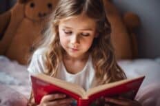 Petite fille couchée sur le ventre dans un lit et lisant un livre à la couverture rouge