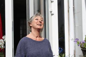 Photo d'Anne Gruwez, à l'extérieur devant ce qui ressemble à une porte vitrée d'une maison, regardant vers le haut en souriant