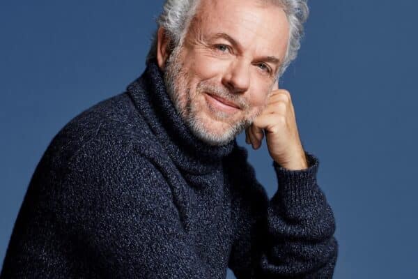 Frédéric Lenoir, la joue posée contre sa main, souriant à la caméra devant un fond bleu.