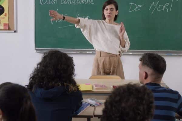 Une institutrice tenant le bras droit, devant des élèves assis sur des bancs dans une classe d'école