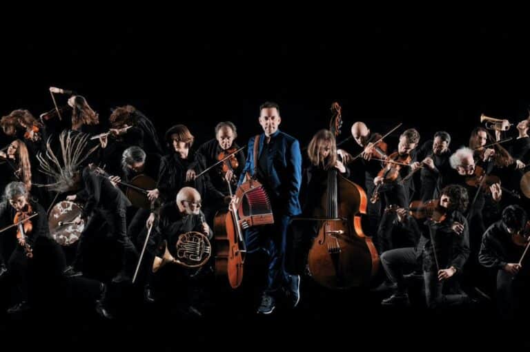 Didier Laloy en costume bleu tenant un accordéon, entouré par d'autres musiciens avec des instruments