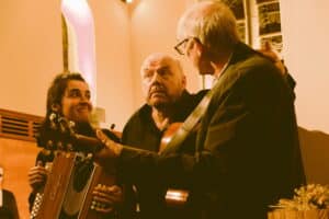 Une femme et deux hommes se tiennent l'un à côté de l'autre, l'un des deux hommes joue de la guitare.