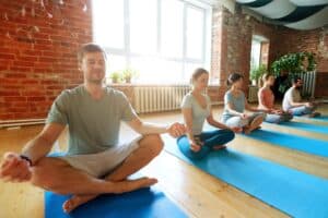 Plusieurs personnes sont assises en tailleur dans une posture de yoga, les yeux fermés, sur des tapis bleus dans une pièce lumineuse