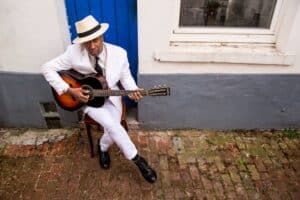 Bai Kamara assis contre un mur blanc et gris, dans un costume blanc, jouant de la guitare