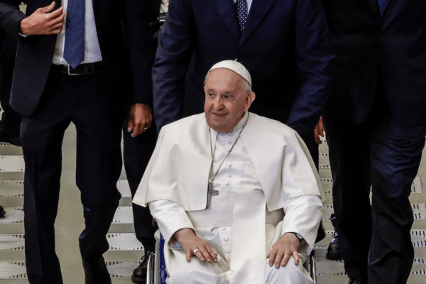 Le pape François vêtu tout de blanc, avec une croix autour du cou, assis sur un fauteuil roulant et des personnes en costume noir marchant derrière lui