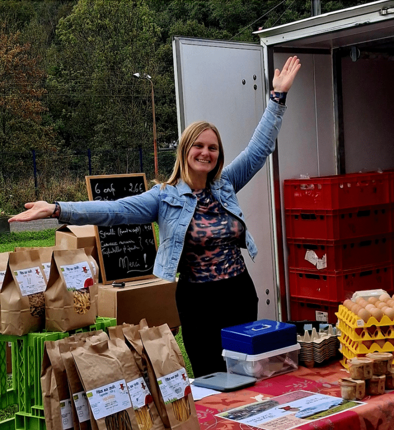 Une femme souriant, les bras vers le haut, derrière une table avec des produits bio dessus