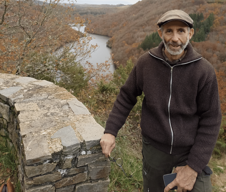 Cassingena Trévédy appuyé sur un muret, avec des arbres et un cours d'eau derrière lui