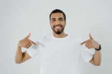 Un homme au t-shirt blanc souriant et se désignant avec les mains