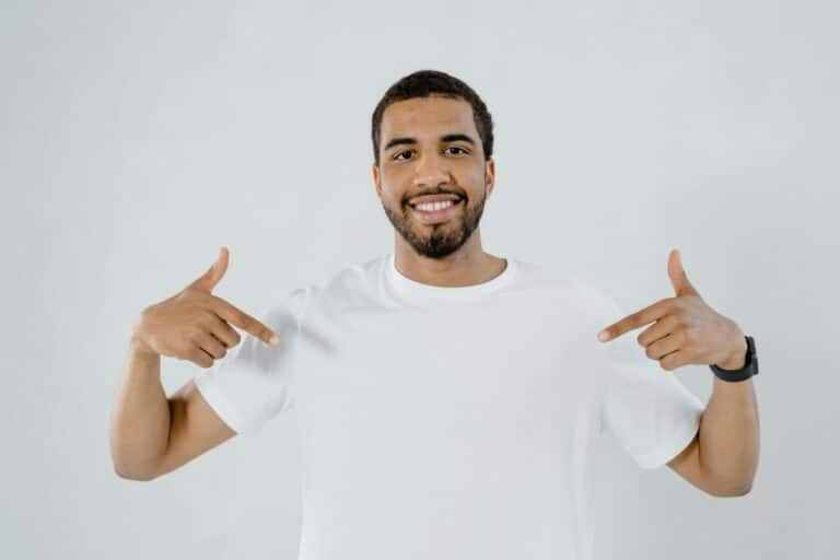 Un homme au t-shirt blanc souriant et se désignant avec les mains