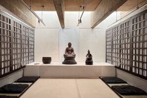 Sur une table blanche, trois objets sont posés. L'objet du milieu représente une figure de Bouddha. Ces objets se trouvent dans une pièce claire