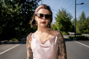 La danseuse Angelina Bruno au milieu d'une route, le ciel est bleu, elle porte une robe rose pale, des lunettes de soleil et des tatouages. Elle a perdu la moitié d'un de ses bras