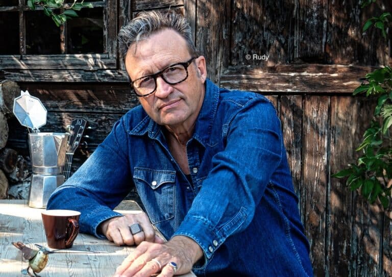Photo de Serge Van Laeken dit "Marka" portant une chemise en jeans bleue. Il est à l'axtérieur accoudé sur une table avec une tasse posée dessus
