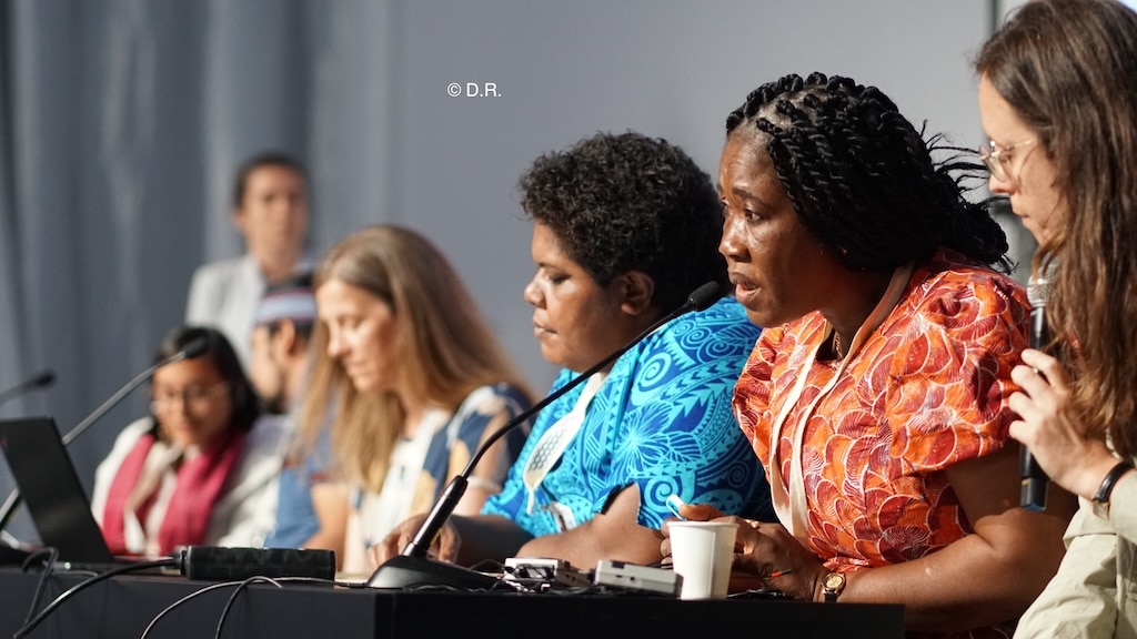 Des femmes alignées et assises les unes à côté des autres dont une a un micro devant elle et parle